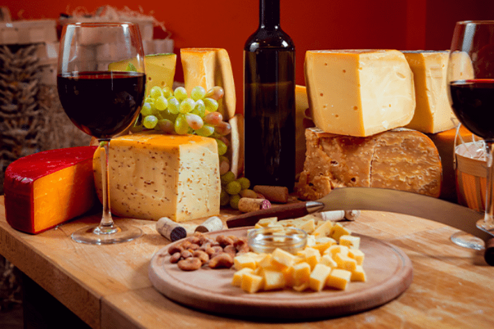 Uma tábua em cima de uma mesa com pedaços de queijo, um cacho de uvas e duas taças de vinho com vinho pela metade. 