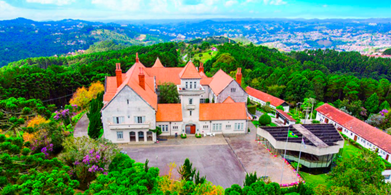 Imagem aérea do Palácio da Boa Vista, uma construção branca com telhados alaranjados, em arquitetura que lembra um castelo. A propriedade fica no centro de uma montanha, cercada de vegetação.