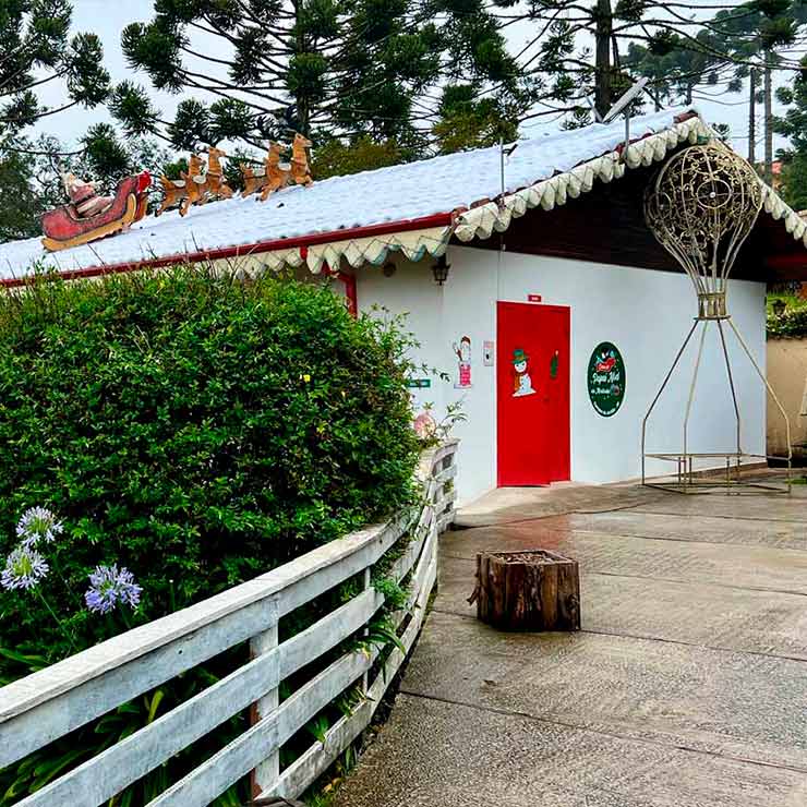 Imagem de uma casa decorada para o Natal, com elementos festivos como um trenó, árvores e neve artificial. A entrada é destacada pela porta vermelha e a vegetação ao redor. Ideal para planejamentos de festas e decoração natalina.